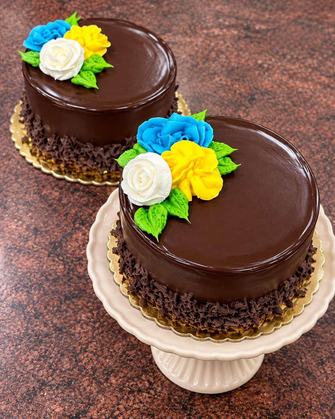 Two round chocolate cakes with glossy glaze and piped blue, yellow, and white flower decorations on a speckled brown countertop. One cake sits on a gold base and the other on a cream cake stand.