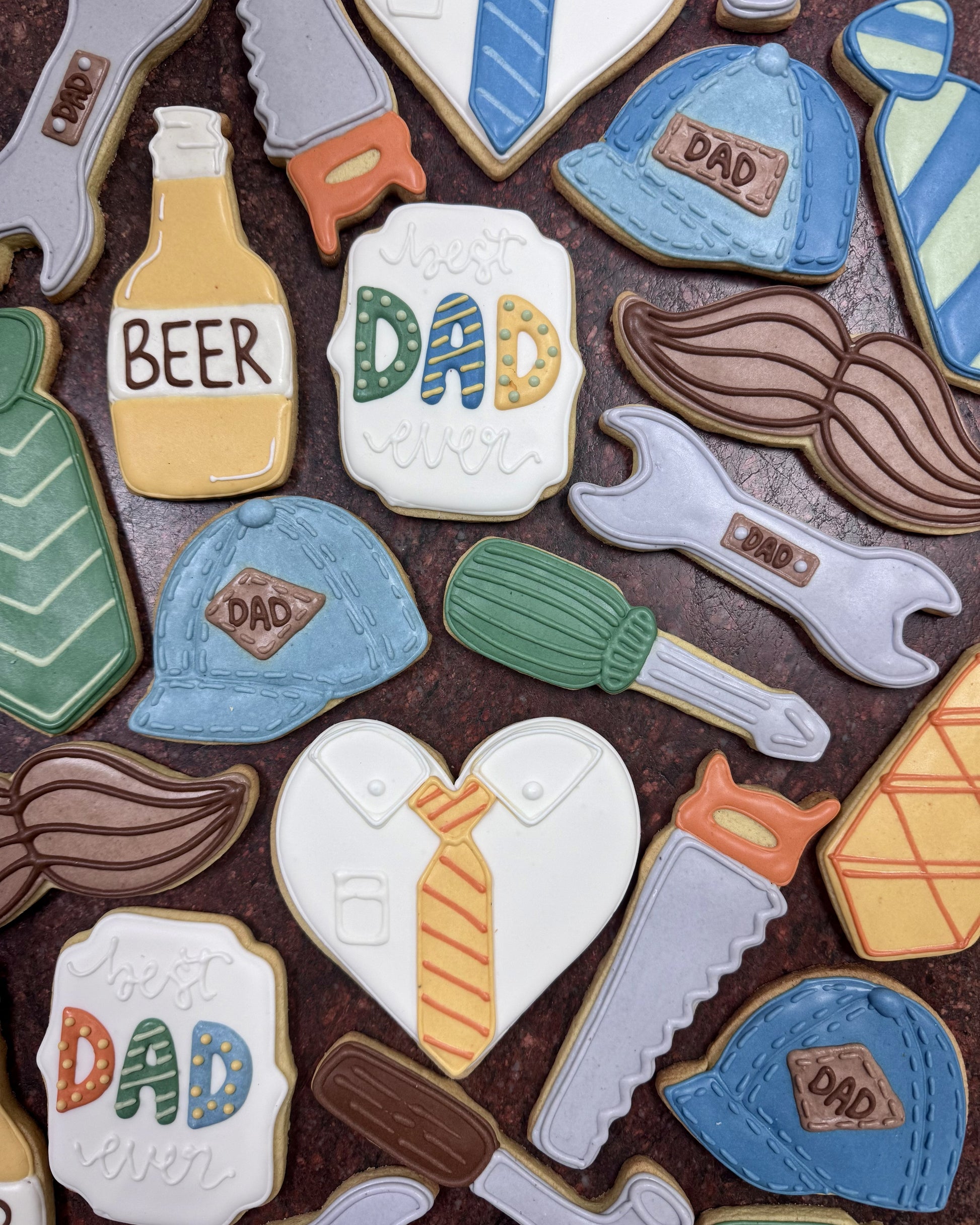Collection of father-themed cookies including tools, beer bottle, and 'DAD' text on a brown background.