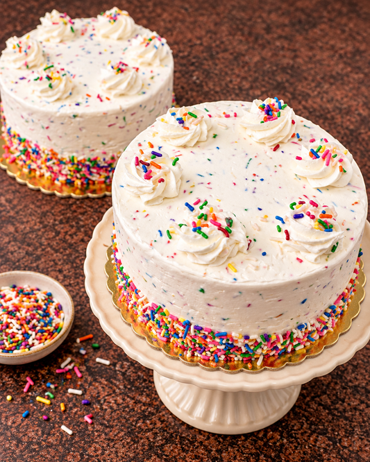 A round white frosted cake topped with piped swirls of whipped cream and colorful sprinkles, displayed on a white pedestal stand on a brown textured surface. A second cake is blurred in the background, and a small dish of sprinkles sits to the left.