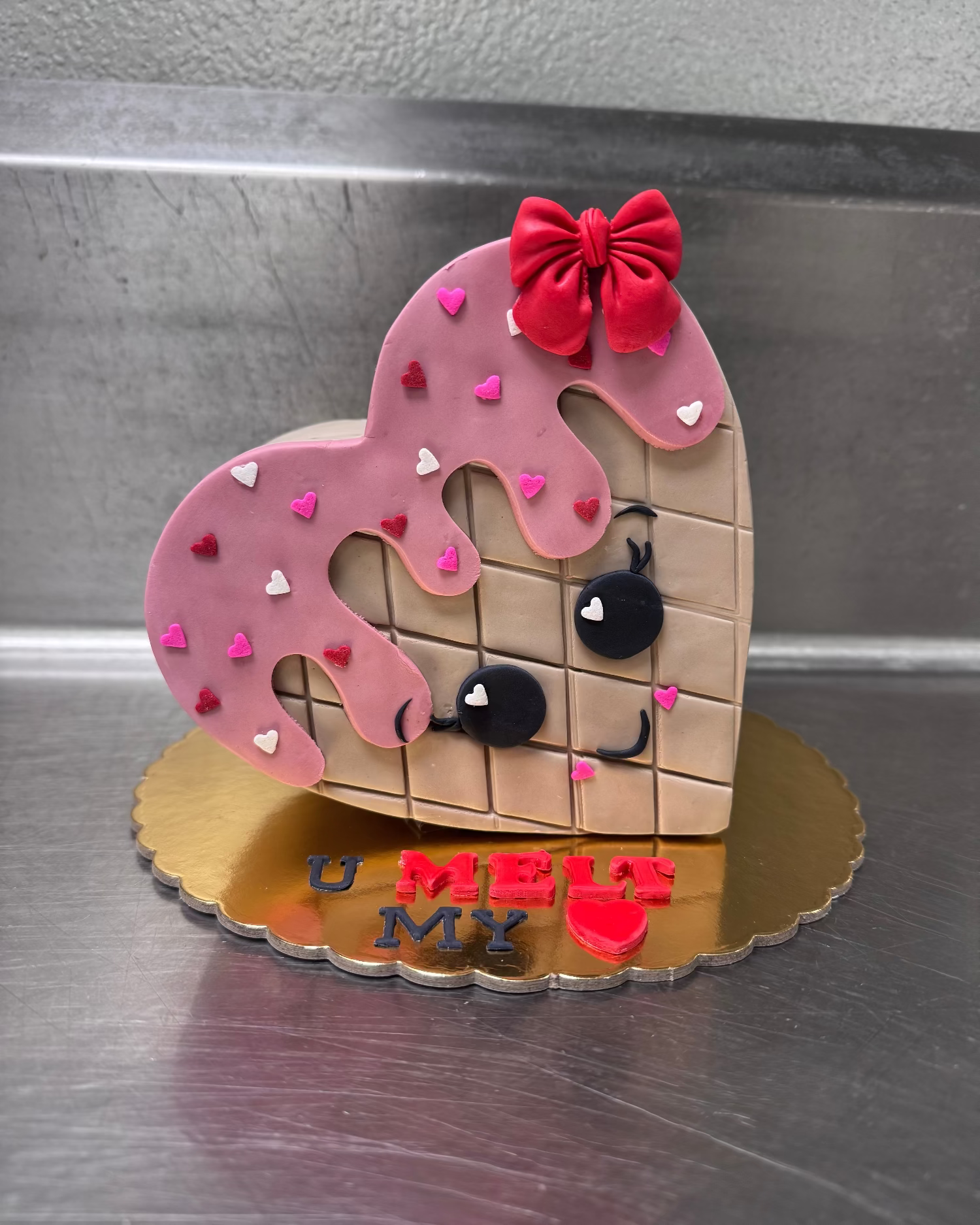 Heart-shaped cookie with a face, pink icing, and red bow on a metallic surface.