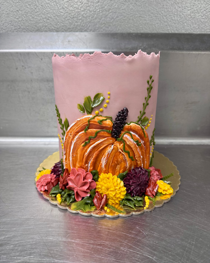 Decorative cake with pink frosting, pumpkin, and flowers on a metallic surface