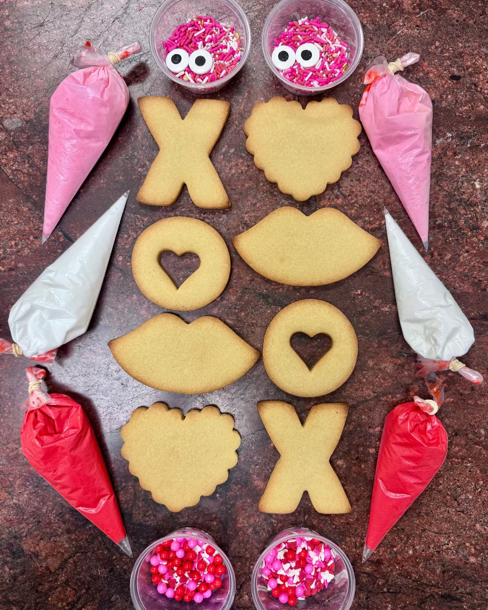 Decorative cookies shaped like lips, hearts, and 'XO' on a brown surface with icing bags and sprinkles.